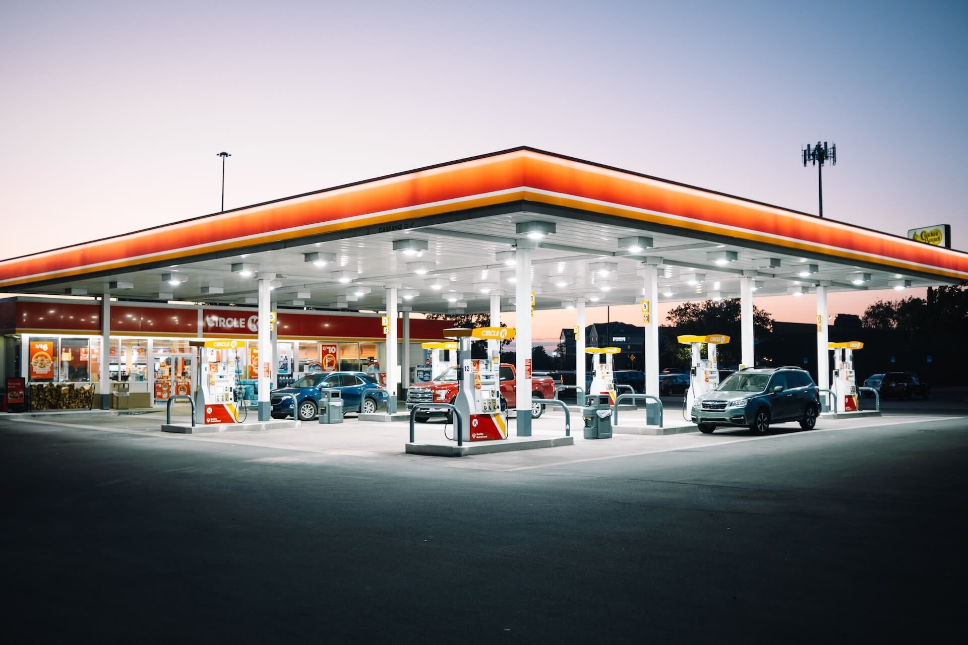 A brightly lit gas station at dusk with cars refueling and a convenience store in the background.