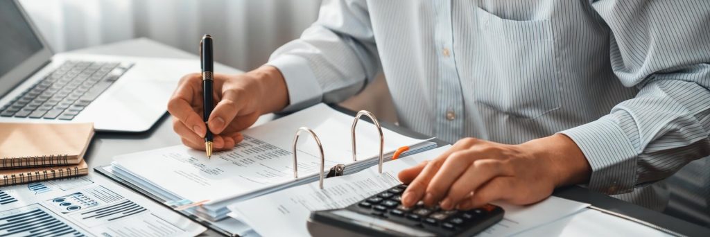 Corporate auditor calculating budget with calculator on his office desk. Dedicated accountant professional of accounting business company analyzing financial document to forecast income. Insight